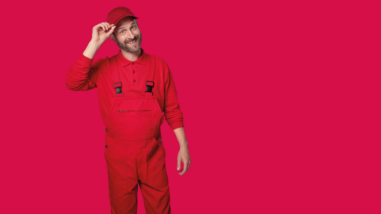 Bärtiger Mann in roter Arbeitskleidung mit Begrüßungsgeste vor neutralem Hintergrund in magenta